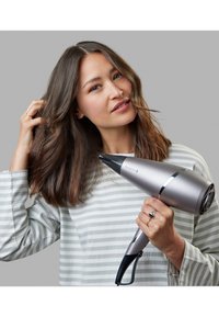 Ασημί πιστολάκι μαλλιών με κομψό σχέδιο, χαρακτηρισμένο από ένα κωνικό ακροφύσιο, μαύρη λαβή και μια γυαλιστερή λωρίδα. Χρήστης στυλιζάρει μακριά, κυματιστά μαλλιά.