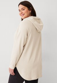 Beige Kapuzenpullover mit langen Ärmeln, mit abgerundetem Saum und weicher Textur. Auf einem Modell aus seitlicher Perspektive präsentiert.