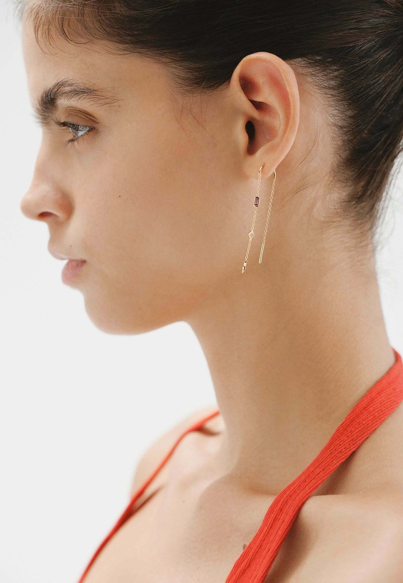 Pendientes de oro colgantes que presentan una cadena delgada, una piedra rectangular púrpura y detalles geométricos intrincados. Modelo mostrando los pendientes de vista lateral.