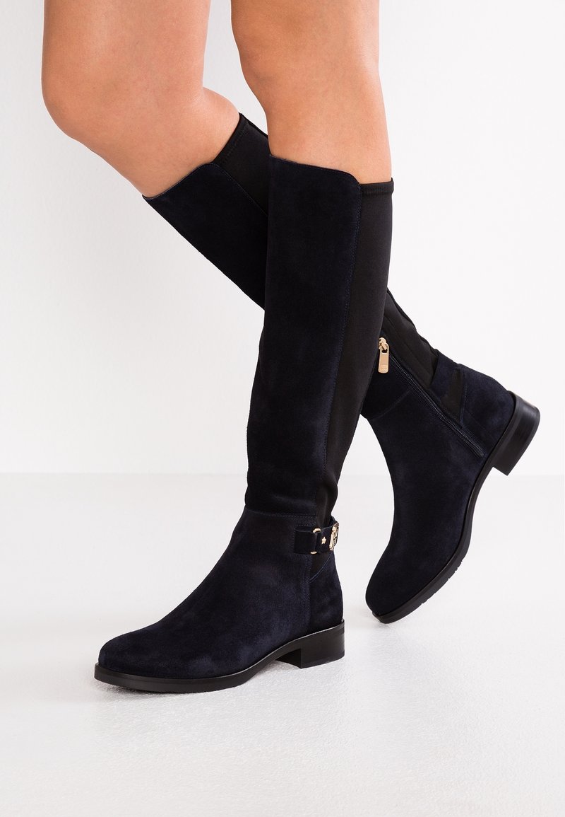 Gambe che indossano stivali neri in suede al ginocchio con cerniera laterale e piccoli dettagli con fibbia, in piedi su un pavimento bianco contro un muro bianco.