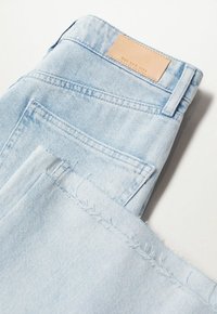 Jeans de mezclilla azul claro con un dobladillo deshilachado, detalles cosidos y una etiqueta de cuero marrón en la cintura trasera. Tejido suave y texturizado.