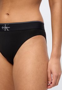 Κοντινό πλάνο ενός ατόμου που φοράει μαύρα εσώρουχα Calvin Klein με το λογότυπο στη μέση, εμφανίζοντας την κοιλιακή περιοχή και το δεξί χέρι να ακουμπά στο μηρό.