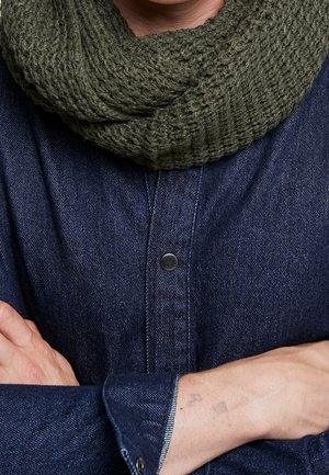 Homme portant une chemise en denim bleu foncé et une écharpe en tricot vert olive, les bras croisés, montrant le poignet et une partie de la main.