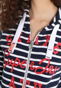 Sudadera con capucha a rayas en azul marino y blanco, con cremallera, que presenta el texto en rojo "SuperDry". Material de algodón, con detalles en cuerda blanca en la capucha.