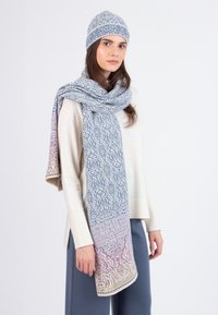 Donna che indossa un maglione crema, pantaloni blu-grigi, e un cappello di maglia e una lunga sciarpa coordinati nei toni di blu, rosa e beige.