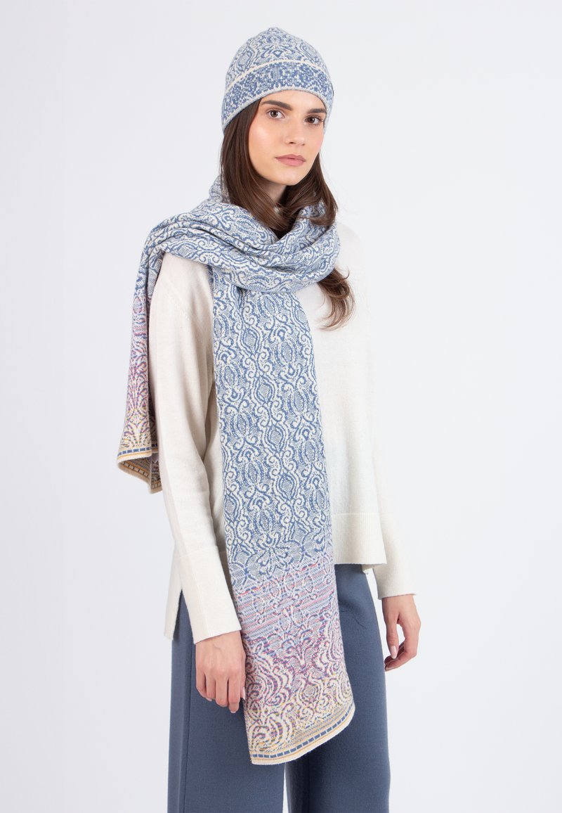 Donna che indossa un maglione crema, pantaloni blu-grigi, e un cappello di maglia e una lunga sciarpa coordinati nei toni di blu, rosa e beige.