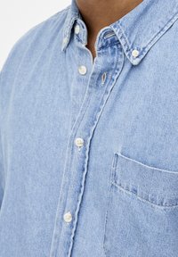 Hellblaues Jeanshemd mit einem Button-Down-Kragen, weißen Knöpfen und einer Brusttasche. Glatte Textur mit dezenten Verwaschungseffekten.