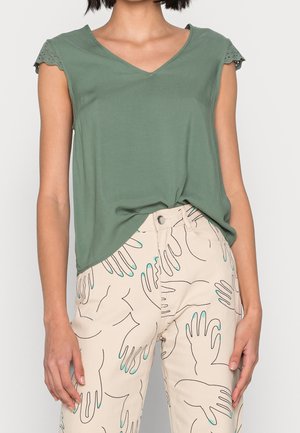 Kobieta ubrana w zielony top z dekoltem w serek, z rękawkami typu eyelet i beżowe spodnie z nadrukiem czarnych konturów dłoni z turkusowymi paznokciami.