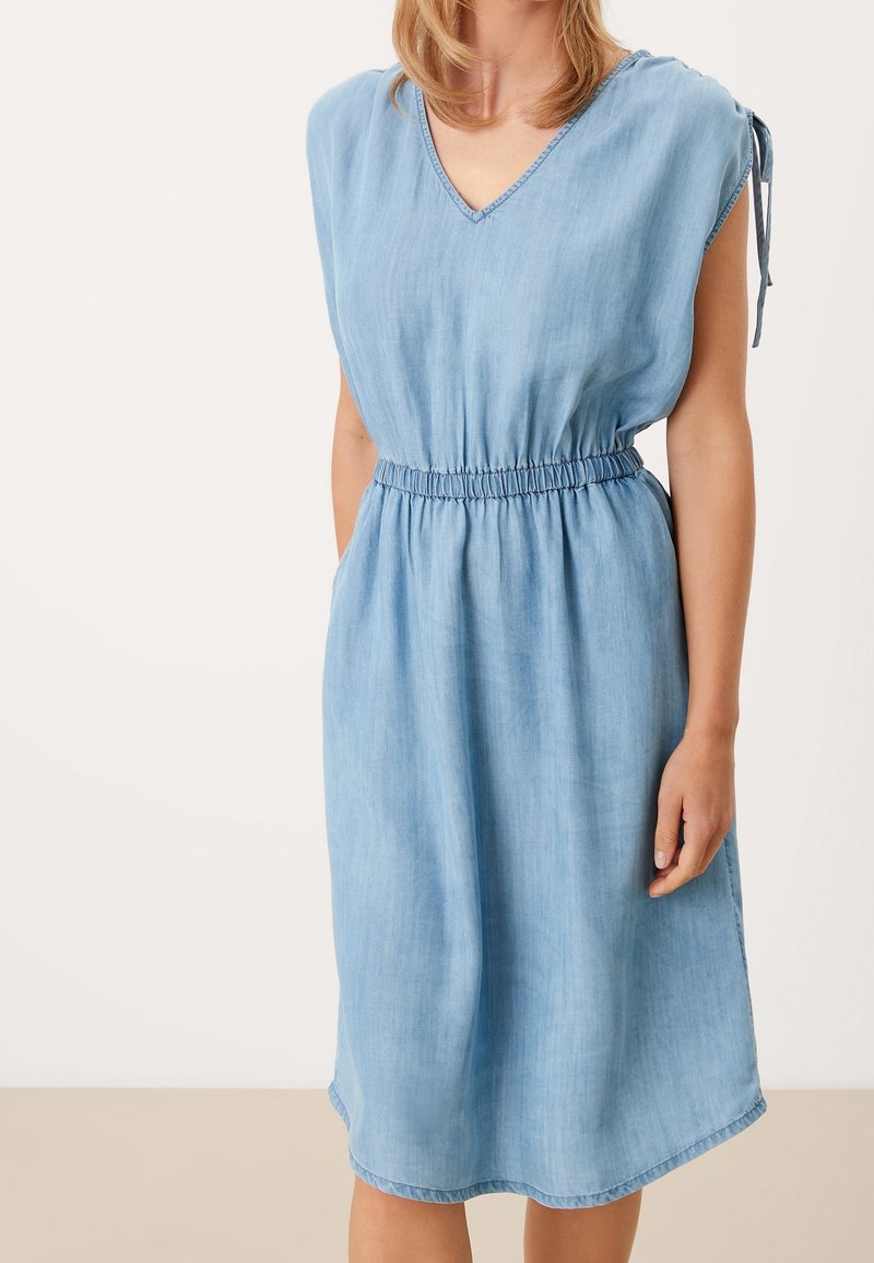 Ljust blå denimklänning med korta capesärmar, V-ringning, elastisk midja och fickor på båda sidor. Slät textur och knälång.