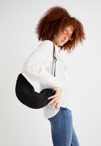 Borsa a tracolla nera a forma di mezzaluna, con un design testurizzato e una tracolla regolabile, tenuta da una persona in camicia bianca.