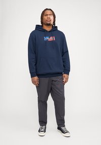 Mørkeblå hettegenser med frontlogo i rødt og hvitt, kombinert med grå bukser og svarte høye sneakers. Ensfarget bakgrunn.