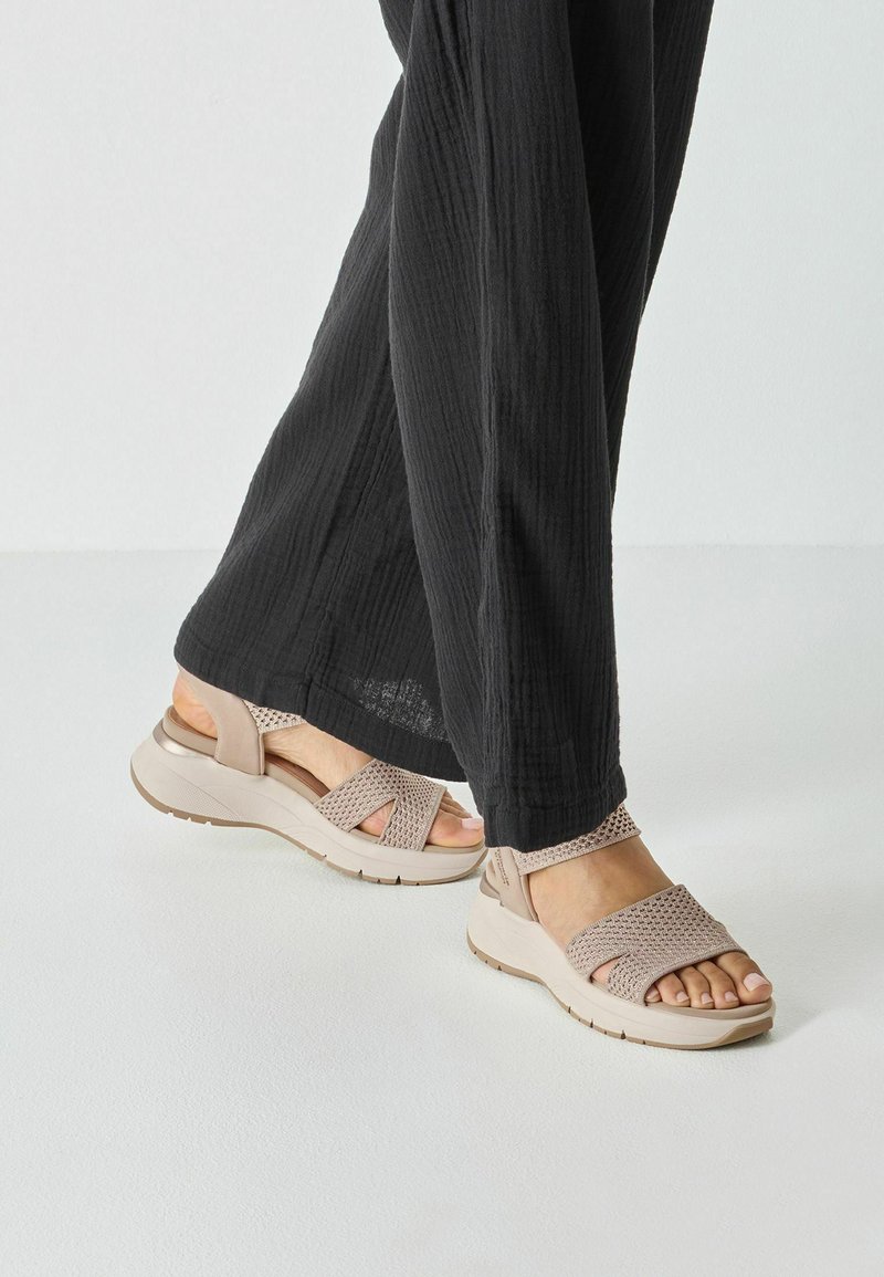 Des sandales beiges avec une tige tissée et des lanières croisées, une semelle rembourrée et une semelle extérieure texturée, associées à un pantalon noir ample.