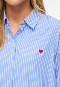 Chemise rayée bleu clair en tissu à texture lisse. Présente un petit cœur rouge brodé sur le côté gauche de la poitrine.