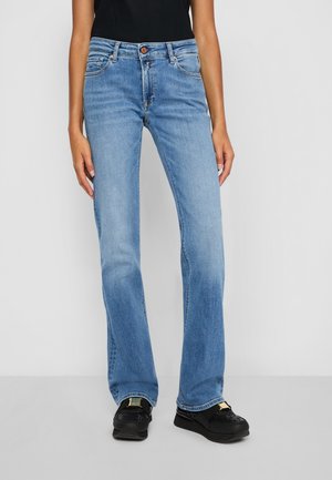 Person iført lyseblå jeans med lige ben, sort top og sorte slip-on sko, stående mod en ensfarvet baggrund.