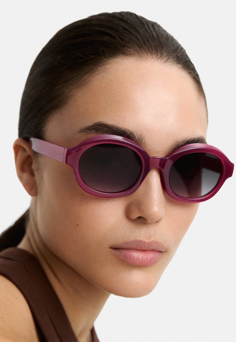 Okrągłe, burgundowe okulary przeciwsłoneczne z ciemnymi szkłami. Grube, błyszczące oprawki i cienkie zauszniki. Zbliżenie na wzór i kolor.