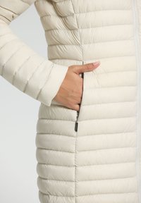 Gevoerde crèmekleurige jas met verticale quilten, een ritszak aan de zijkant en een gladde, glanzende textuur. Voorkant met ritsluiting.