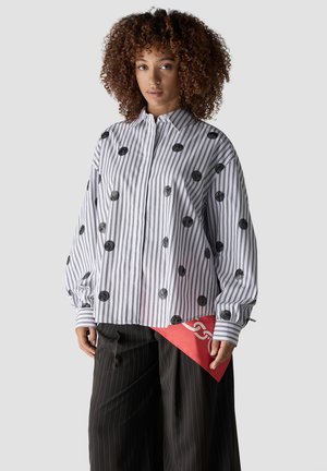 Frau mit lockigem Haar trägt eine weiße Bluse mit schwarzen Streifen und Punkten, schwarze weite Nadelstreifenhose und hält eine rote Clutch-Tasche.