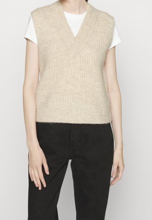 Osoba ubrana w beżową dzianinową kamizelkę z dekoltem w serek, na białej koszuli z krótkim rękawem i czarnych spodniach, stojąca na jednolitym tle.