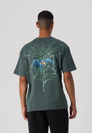 Mann trägt dunkelgrünes Nike SB T-Shirt mit grünem Spinnennetz und blauem Spinnengrafik auf der Rückseite, steht vor einfarbigem grauem Hintergrund.