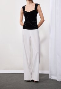 Top sin mangas de terciopelo negro con escote cuadrado y detalle fruncido, combinado con pantalones de pierna ancha blancos y sandalias de tacón negras.