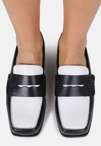 Mocasines de cuero en blanco y negro con punta cuadrada. Presentan una amplia cinta negra con diseño de corte y costuras en contraste a lo largo de los bordes.
