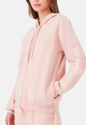 Haut à capuche zippé rose en tissu doux. Il dispose d'une capuche avec cordon de serrage, de poches avant et d'un texte logo blanc sur la manche gauche.