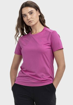 Woman with long brown hair wearing a bright pink short-sleeve shirt and black pants, standing with hands in pockets against a plain background.