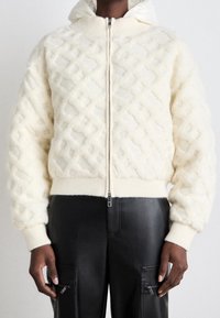 Krämfärgad fleecejacka med strukturerad diamantmönster, dragkedja, ribbade ärmslut och nederkant. Kombineras med svarta läderbyxor med fickor.