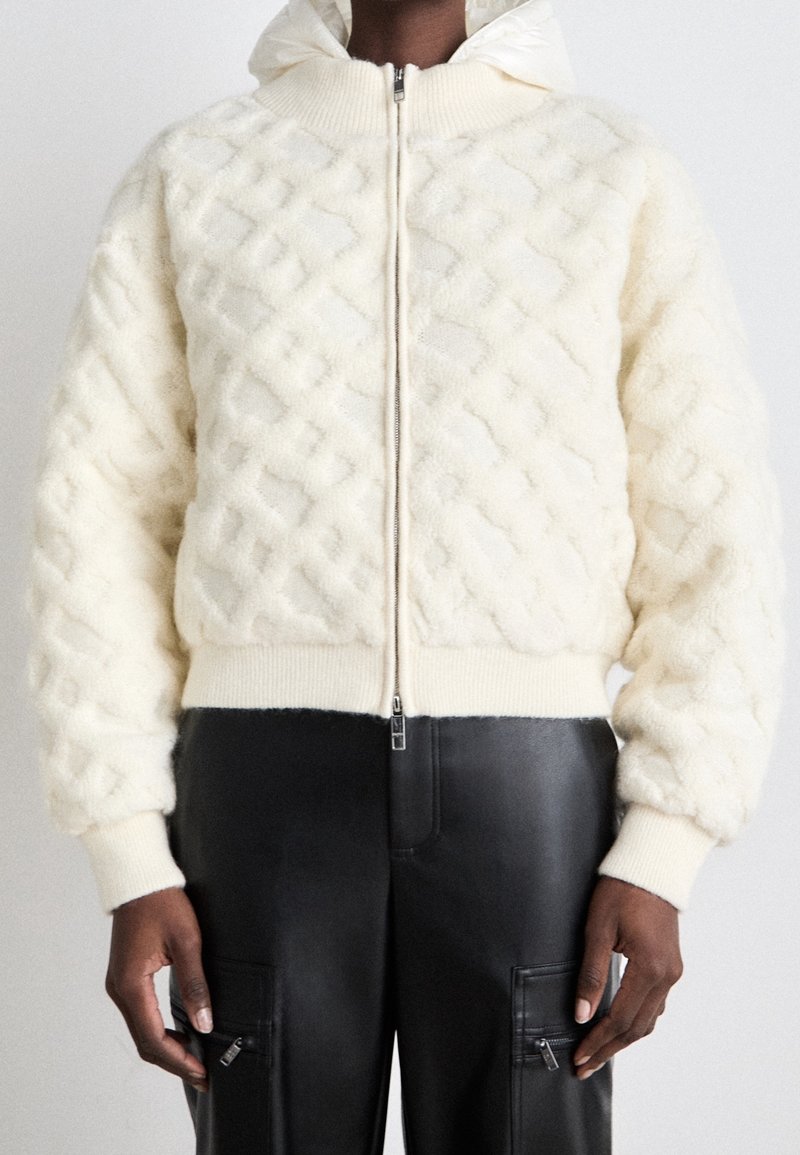 Krämfärgad fleecejacka med strukturerad diamantmönster, dragkedja, ribbade ärmslut och nederkant. Kombineras med svarta läderbyxor med fickor.