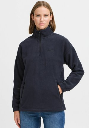 Fleece pullover in dark navy, featuring a half-zip, zippered side pockets, and a logo patch on the chest. Soft texture and loose fit.