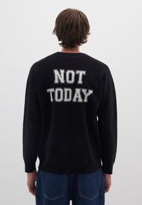 Felpa nera oversize con "NOT TODAY" in grande testo bianco sulla schiena. Tessuto morbido, polsini a costine e orlo, abbinata a pantaloni di jeans blu.