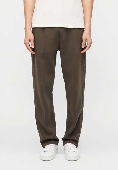 Personne portant un pantalon marron à jambes droites et des baskets blanches, debout devant un fond clair uni, les mains relâchées le long du corps.