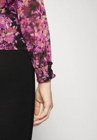 Blusa a maniche lunghe con motivo floreale nei toni di viola, rosa e nero, con polsini arricciati e volant. Abbinata a una gonna nera.