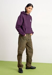 Purple hoodie with a triangular logo, paired with olive green, loose-fitting pants and black footwear. Simple background with green floor.