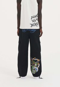 Schwarze Cargo-Hosen mit grafischer Stickerei, die Schädel- und Adlerdesigns zeigen, kombiniert mit einem weißen Tanktop, auf dem "PUNK NOT DEAD" steht.