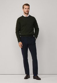 Maglione verde scuro a maglia con un motivo a trecce, indossato sopra una camicia bianca con colletto. Abbinato a pantaloni blu scuro e scarpe eleganti scure.