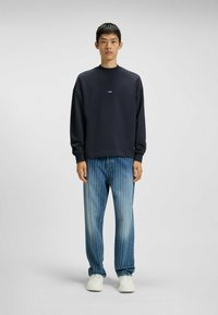 Marinblå sweatshirt med rund hals och liten etikett på bröstet, tillsammans med ljusblå randiga jeans och vita sneakers.