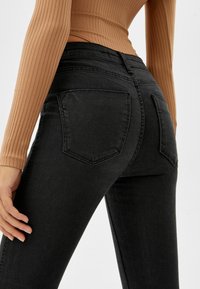 Jeans de mezclilla negra con corte ajustado, que cuentan con dos bolsillos traseros, una textura suave y una cinturilla de altura media. Bordes recortados visibles.
