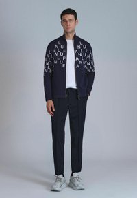 Marine blauwe zip-up jas met grote witte letters, gecombineerd met donkere broeken en lichte sneakers. Glad fabric en sportief ontwerp.