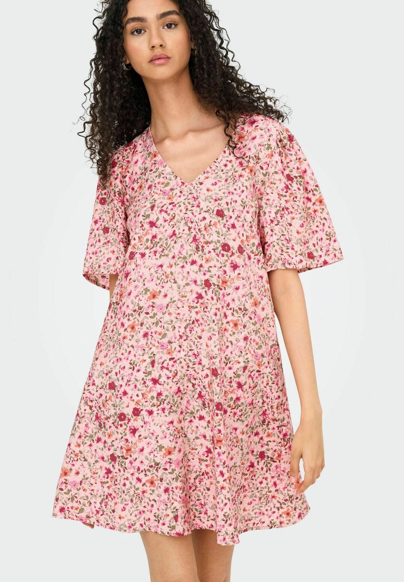 Ziedu kleita gaiši rozā audumā, ar V-veida kakla izgriezumu, īsām pūkainām piedurknēm un izplestu siluetu, rotāta ar mazu ziedu rakstu.