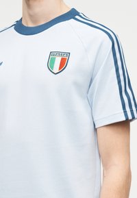 Ανοιχτό μπλε αθλητική μπλούζα με σκούρο μπλε γιακά και ρίγες, σήμα με το τρικολόρ της Ιταλίας στο αριστερό στήθος.