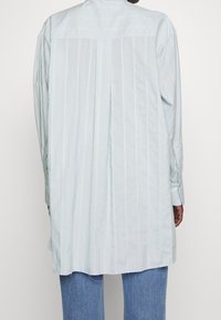 Osoba v světle modré pruhované dlouhé košili s centrálním skladem na zádech, doplněná modrými džíny, otočená zády k fotoaparátu.