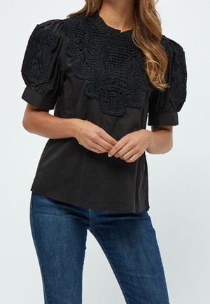 Femme portant un chemisier noir avec des motifs en dentelle complexes sur les manches bouffantes et la poitrine, associé à un jean bleu, les mains délicatement jointes.