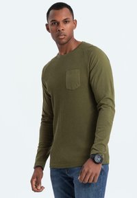 Hombre vistiendo una camisa de manga larga verde oliva con bolsillo en el pecho y jeans azules, de pie con una expresión neutra sobre un fondo blanco.