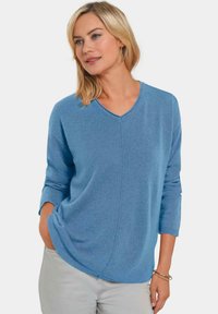 Blauer V-Ausschnitt-Pullover mit strukturierter Oberfläche, Dreiviertelärmeln und diagonaler Naht. Kombiniert mit hellen Hosen.