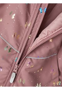 Nærbillede af en pink jakke med frontlynlås, der har metalliske sommerfugle og reflekterende pipingdetaljer på stoffet.