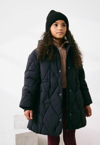 Mustast tikandiga täidetud talvejope kapuutsiga ja nööpidega, millel on sisemine tõmblukuga krae, kantud pruunika topi ja burgundi värvi pükste kohal.