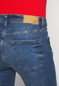 Vaqueros de mezclilla en azul oscuro, con una textura suave, diseño de cinco bolsillos y una etiqueta de cuero marrón en la parte trasera de la cintura con el logotipo.