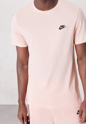 Mężczyzna ubrany w jasnoróżową koszulkę Nike i pasujące krótkie spodenki z czarnymi logo Nike na klatce piersiowej i udzie, stojący na jednolitym tle.