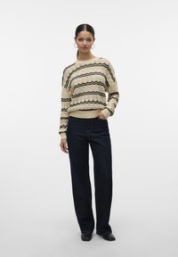 Maglione lavorato a maglia beige con strisce ondulate nere, scollatura rotonda, polsini a coste, abbinato a jeans a gamba larga blu scuro e scarpe nere.